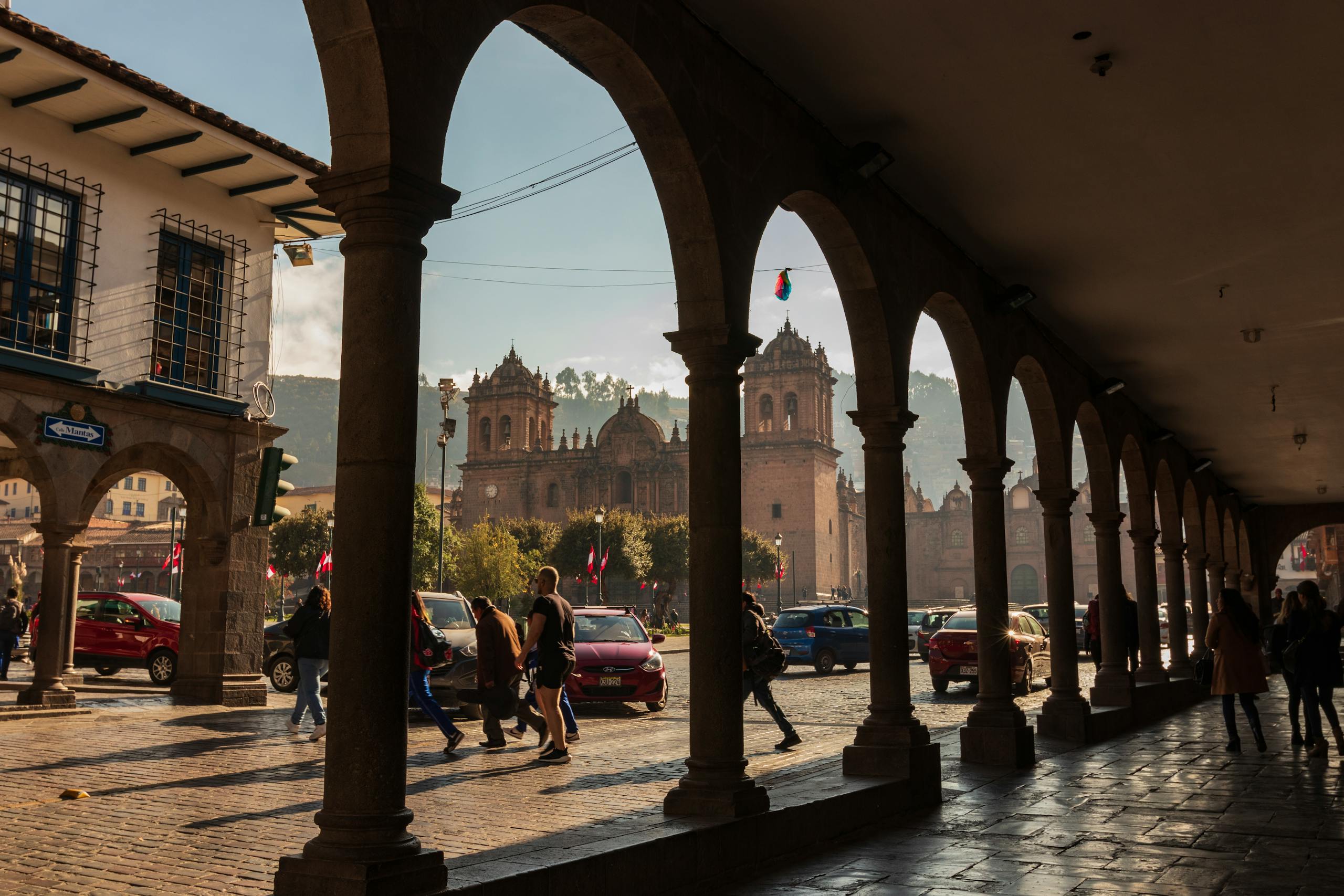 Scenic view of Cusco's cathedral framed by colonnades, capturing urban life with pedestrians and cars.